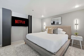 A modern bedroom with a large bed, a wall-mounted TV displaying the Netflix logo, a bedside table with a lamp, and a mirror. - Explore Gaslamp from Chic Loft +Balcony City Views (San Diego)
