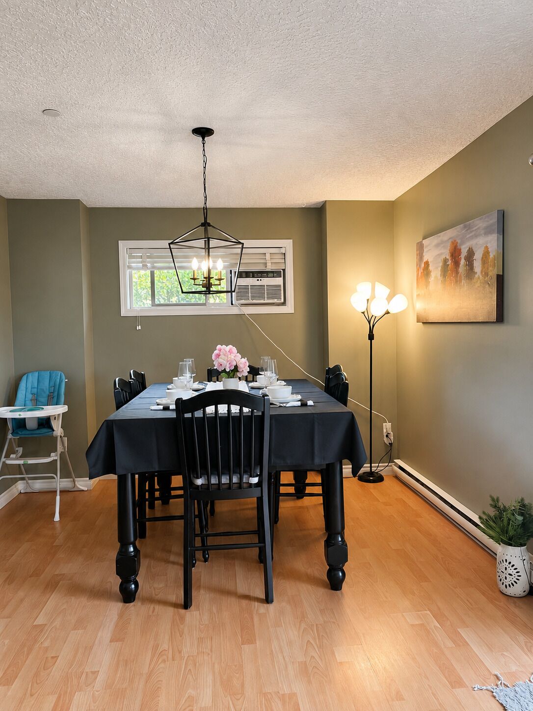 A dining room with a table set for four, a blue high chair, a floor lamp, and a painting on the wall.