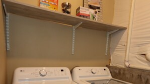 Zona de lavandería con dos lavadoras de carga frontal blancas y un estante superior con una caja de toallas de secadora y un recipiente.