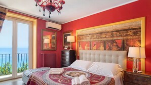 A hotel room with a large bed, a view of the sea, a chandelier, and a painting on the wall.