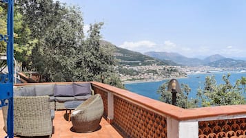 A terrace with wicker furniture, a view of a coastal city, and a mountainous backdrop.