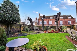 A residential garden with a brick house, a lawn, potted plants, and outdoor furniture.