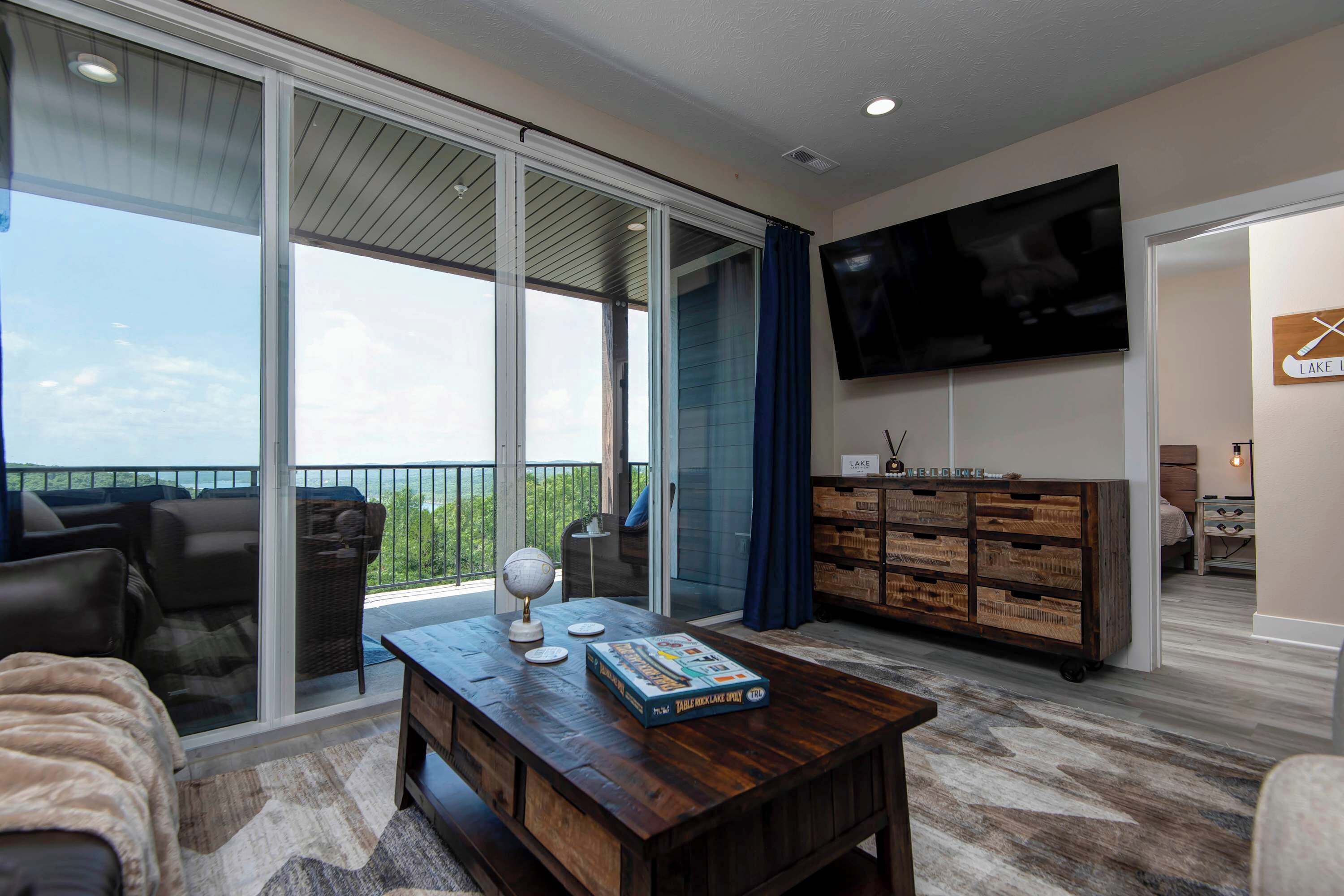 A spacious living area with a large sliding glass door leading to a balcony, a wooden coffee table with a board game, a flat-screen TV mounted on the wall, and a rustic wooden cabinet.