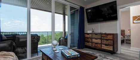 A spacious living area with a large sliding glass door leading to a balcony, a wooden coffee table with a board game, a flat-screen TV mounted on the wall, and a rustic wooden cabinet.