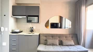 Uma cozinha compacta com micro-ondas, fogão e pia. Um sofá com almofadas está posicionado contra a parede, e um espelho grande reflete parte do quarto.