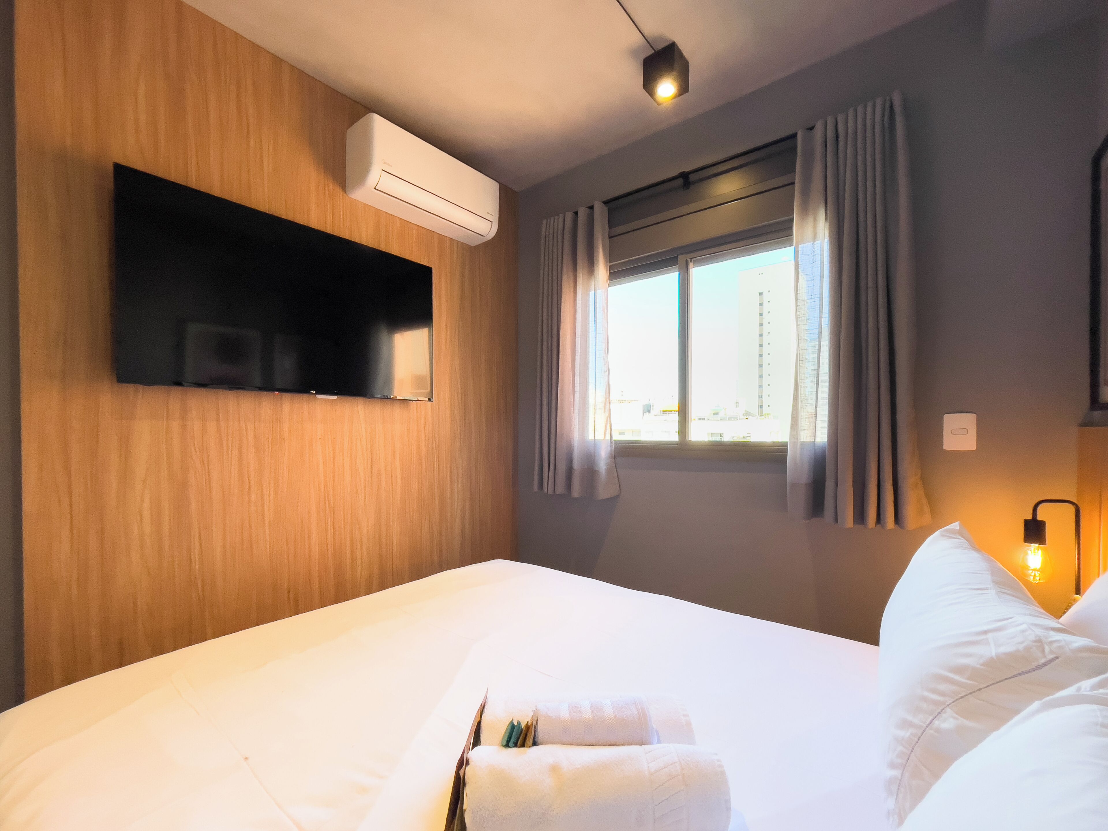 A hotel room with a bed, an air conditioning unit, a mounted television, a window with curtains, and a bedside lamp.