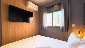 A hotel room with a bed, an air conditioning unit, a mounted television, a window with curtains, and a bedside lamp.