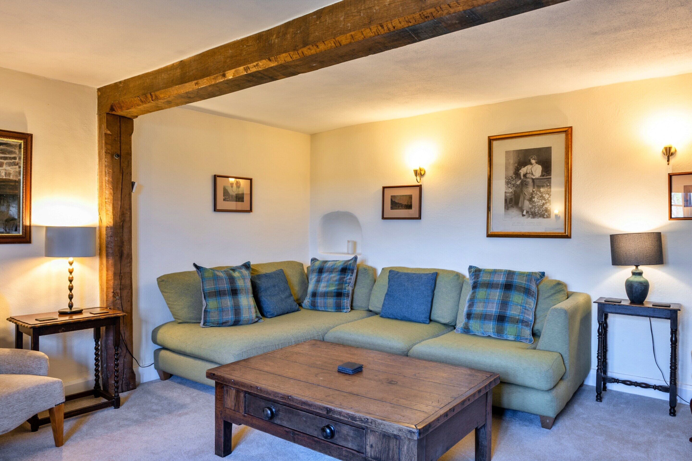 A cozy living room with a wooden beam ceiling, a green sofa with plaid cushions, a wooden coffee table, and framed pictures on the walls.