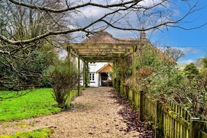 A pathway leading to a traditional house with a thatched roof, surrounded by greenery and a wooden fence. - Luxury Cotswolds Cottage for 7 (Okehampton)