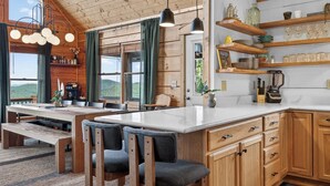 A wooden cabin interior with a dining area, kitchen, and shelves with glassware. - Ski-Out to Hatley Pointe • Alpine Slide • Rec Room (Mars Hill)