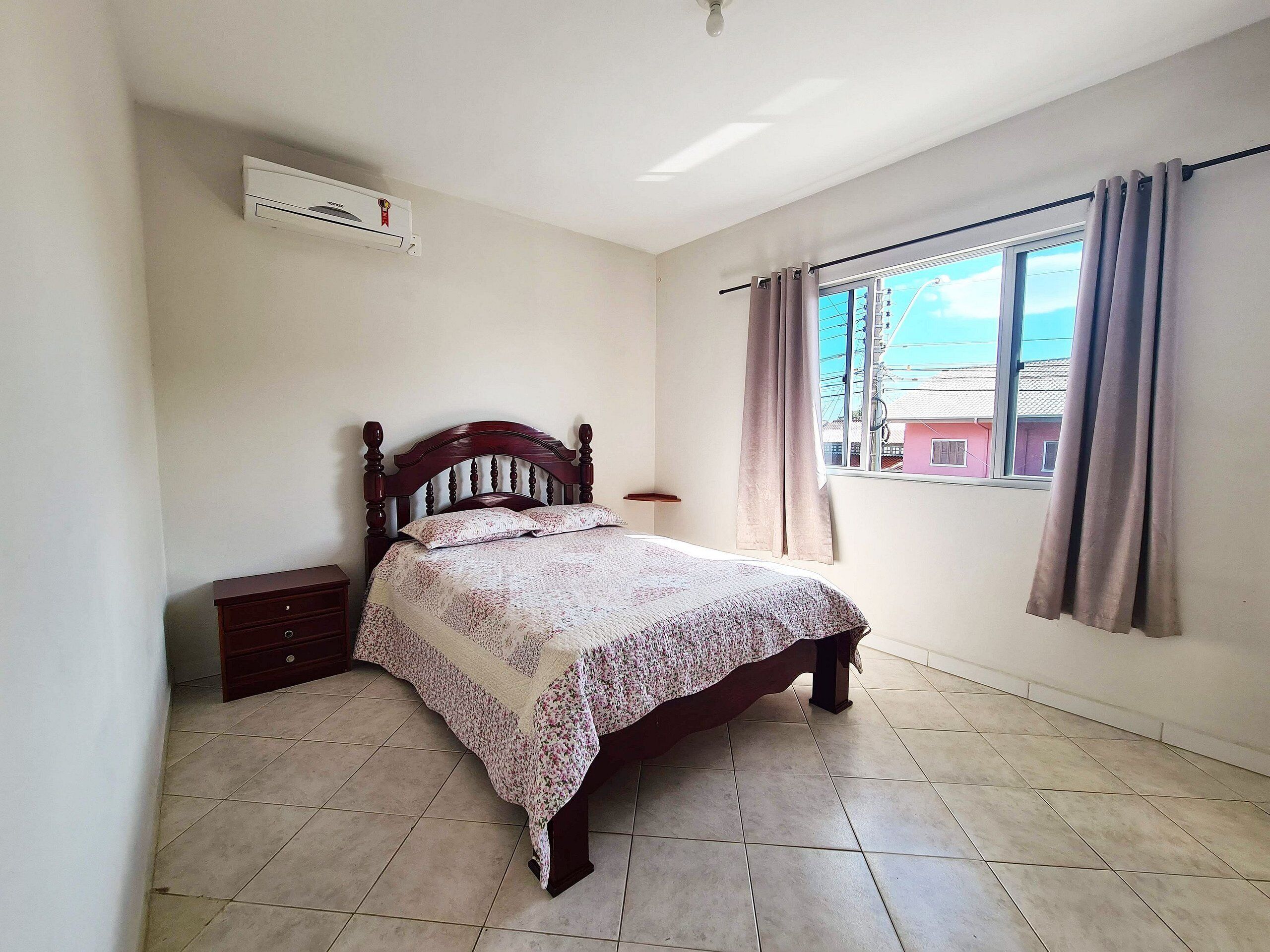 A bedroom with a bed, a nightstand, a window with curtains, and an air conditioning unit.