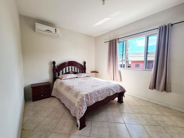 A bedroom with a bed, a nightstand, a window with curtains, and an air conditioning unit. - Canto Grande by LikeHome (Bombinhas)