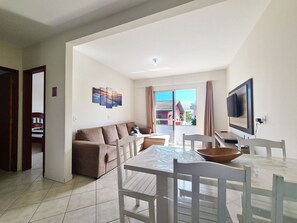 A living room with a sofa, a dining table with chairs, a TV mounted on the wall, and a view through a glass door. - Canto Grande by LikeHome (Bombinhas)