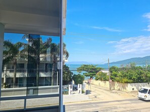 A view from a window overlooking a coastal road with a building and a clear blue sky. - Canto Grande by LikeHome (Bombinhas)