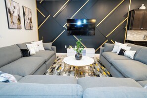 Un salon moderne avec deux canapés gris, une table basse blanche et un téléviseur à écran plat fixé à un mur noir agrémenté de détails dorés.