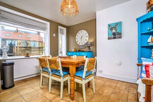 Un comedor con mesa y sillas de madera, un reloj de pared, un aparador azul y una ventana con vista a una casa.