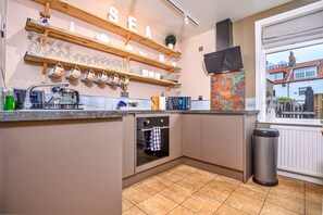 A kitchen with a built-in oven, a sink, and a window overlooking a residential area. - Whitby Coastal Cottage (Whitby)