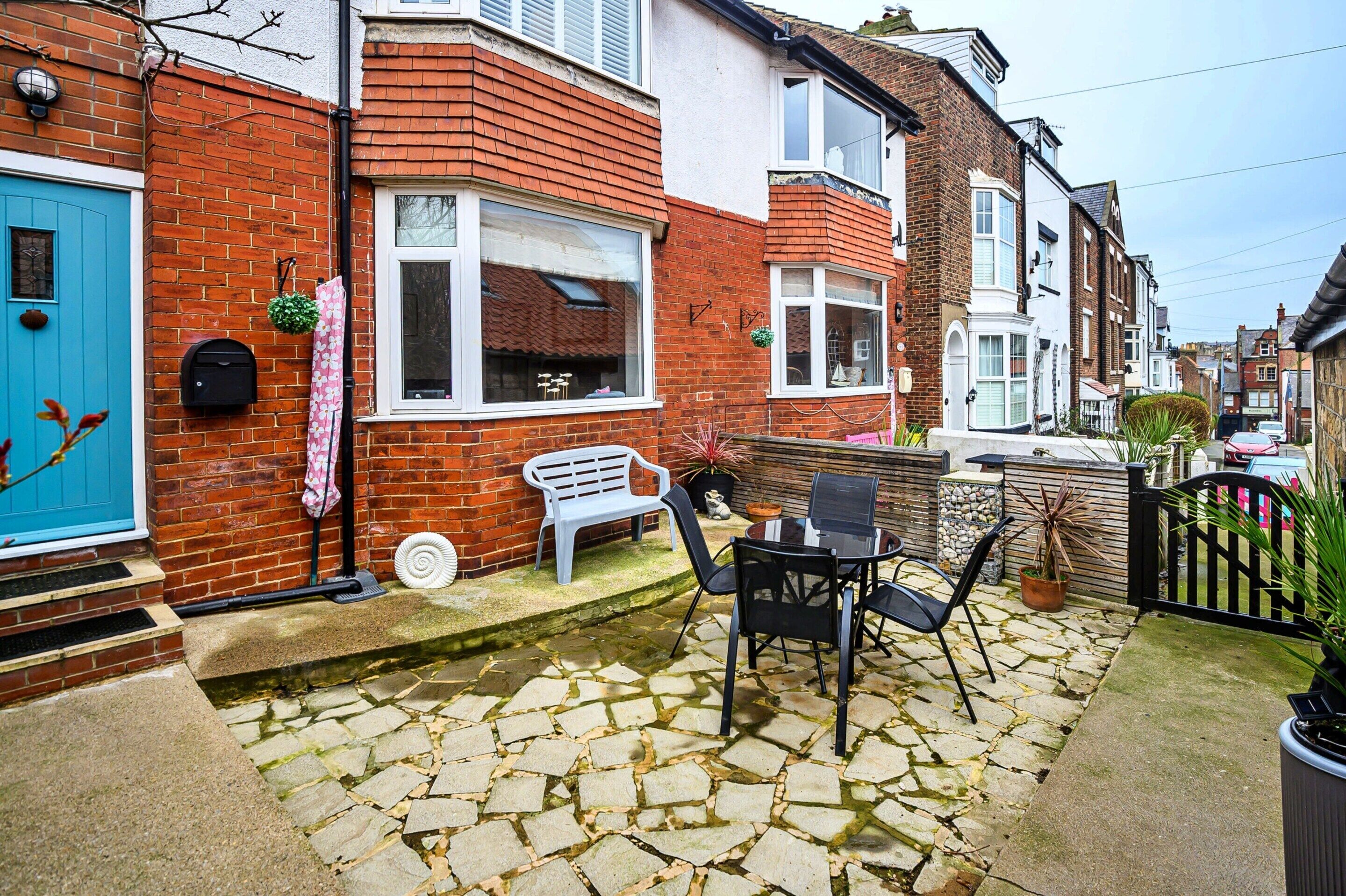 A residential property with a brick facade, a blue door, a small garden with a table and chairs, and a cobblestone pathway.