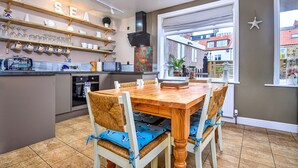 A kitchen with a wooden table and chairs, a built-in oven, and a shelf with glassware. - Whitby Coastal Cottage (Whitby)