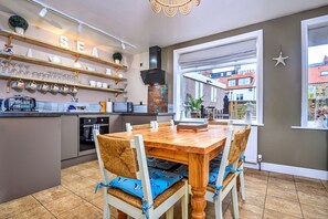 A kitchen with a wooden table and chairs, a built-in oven, and a shelf with glassware. - Whitby Coastal Cottage (Whitby)