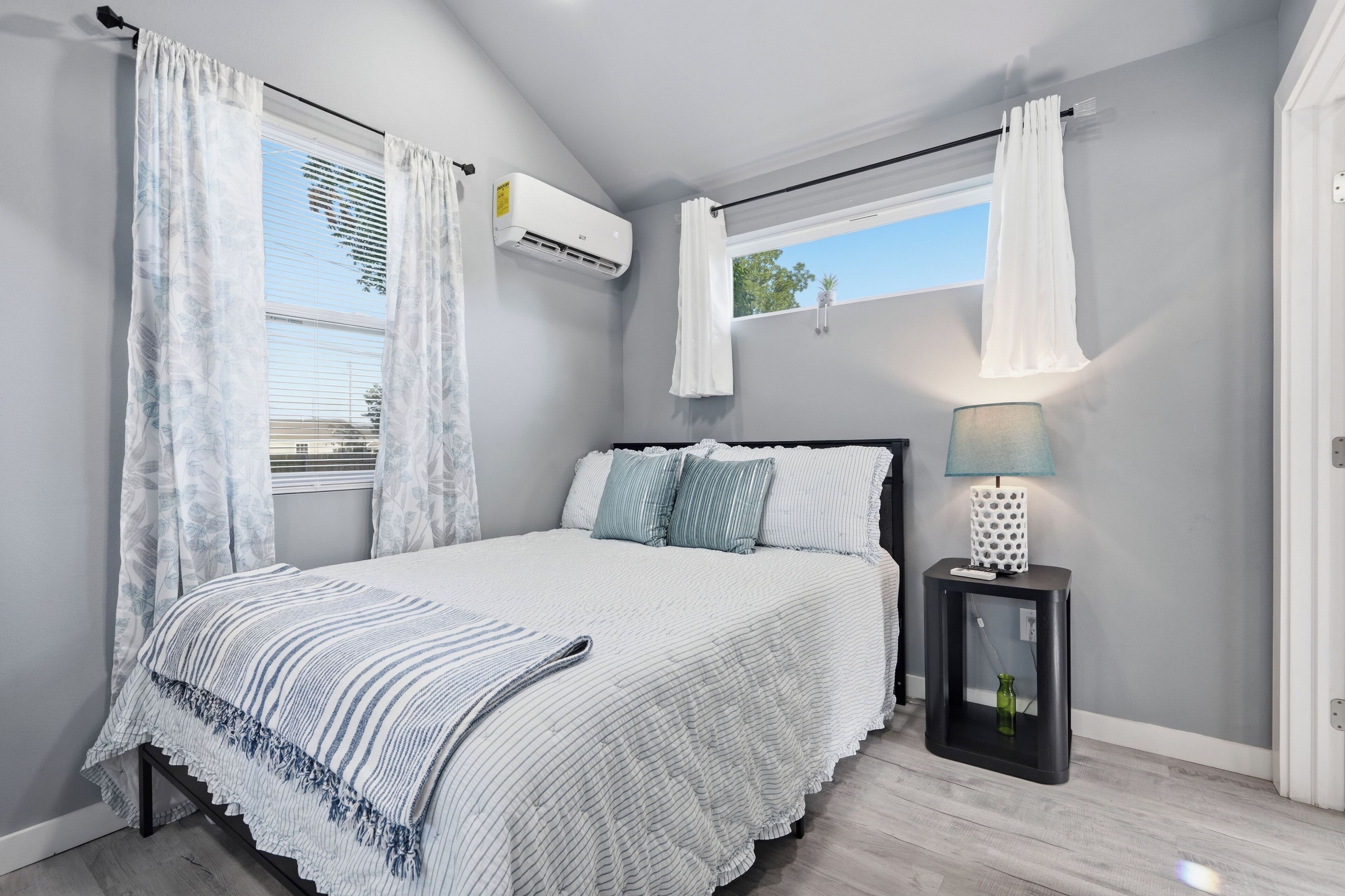 A bedroom with a bed, a nightstand, a lamp, an air conditioning unit, and a window with curtains.