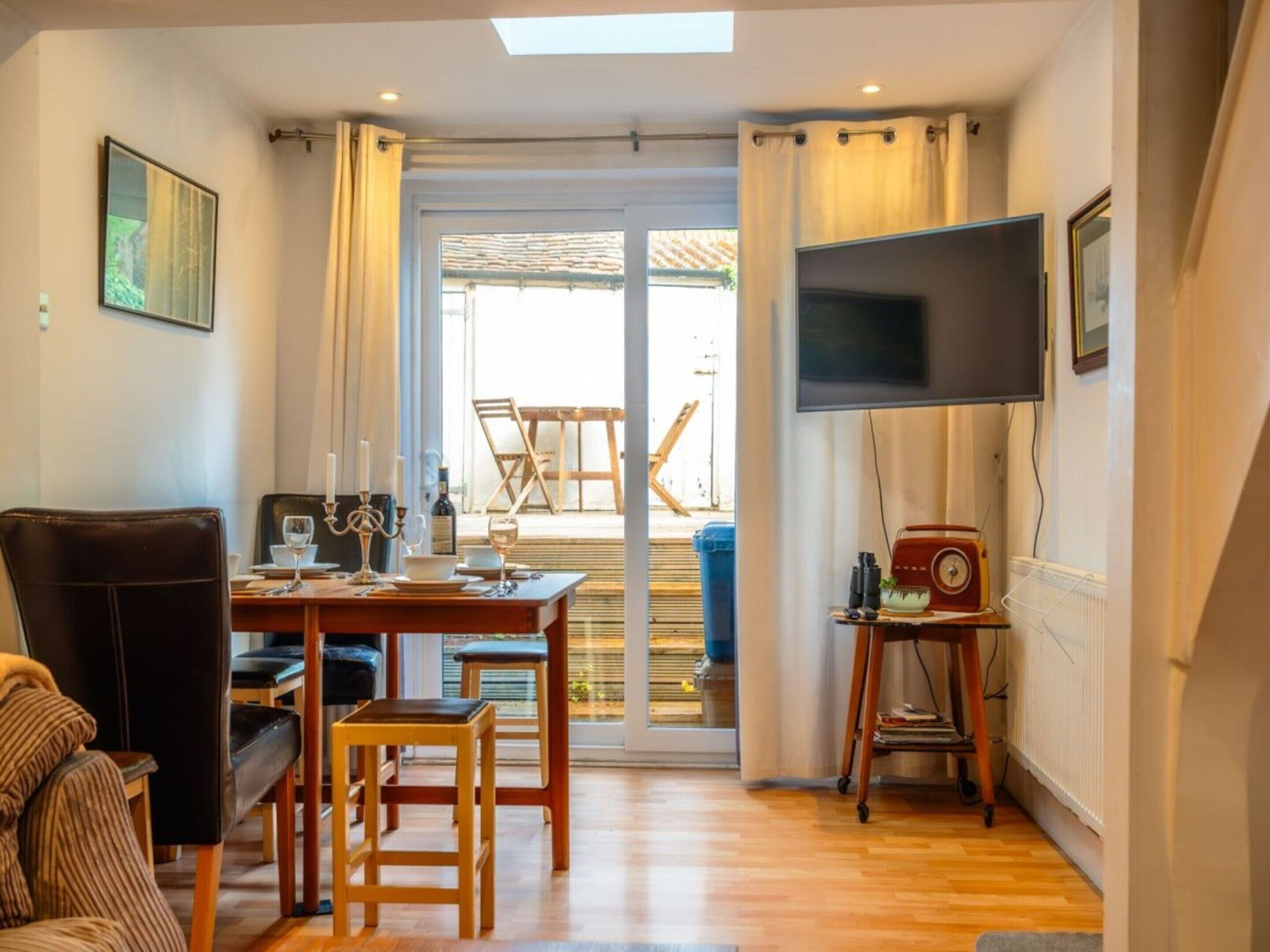 A room with a dining table set for two, a television mounted on the wall, and a window with curtains.