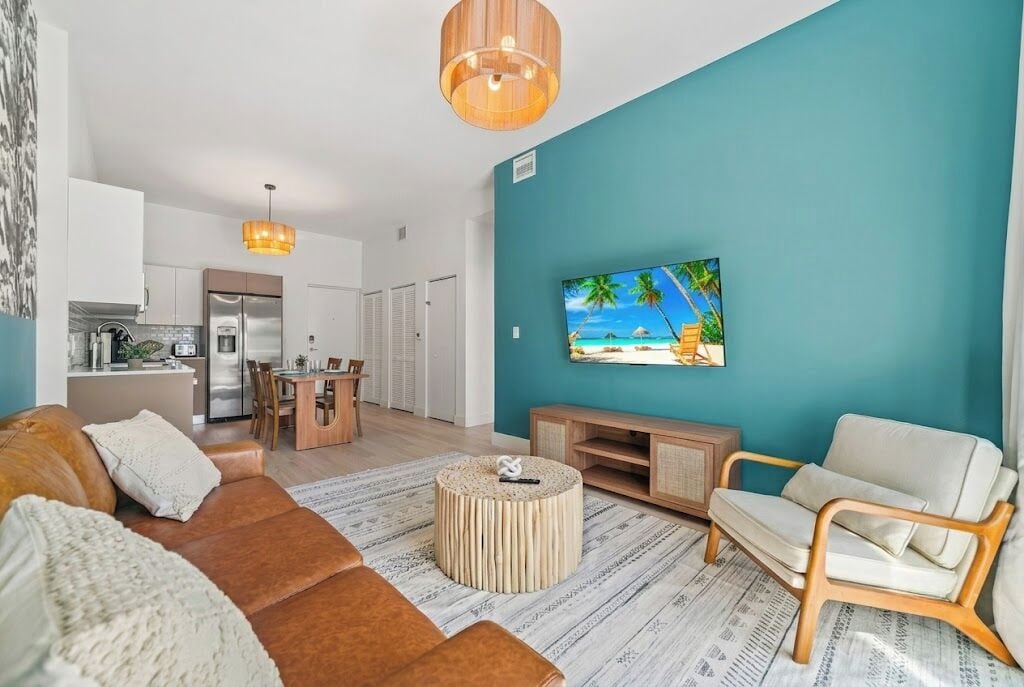 Een moderne woonkamer met een turquoise accentmuur, een flatscreen televisie, een bruine leren bank, een houten salontafel en een eethoek met een keuken op de achtergrond.