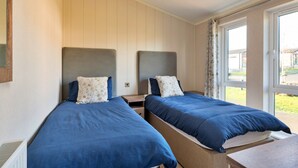 Quarto com duas camas, cada uma com cabeceira e travesseiros, uma pequena mesa de cabeceira de madeira e uma janela com cortinas.