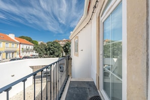 A balcony with a metal railing, a glass door, and a view of the street and buildings. - GuestReady - Riverside Retreat (Alcochete)