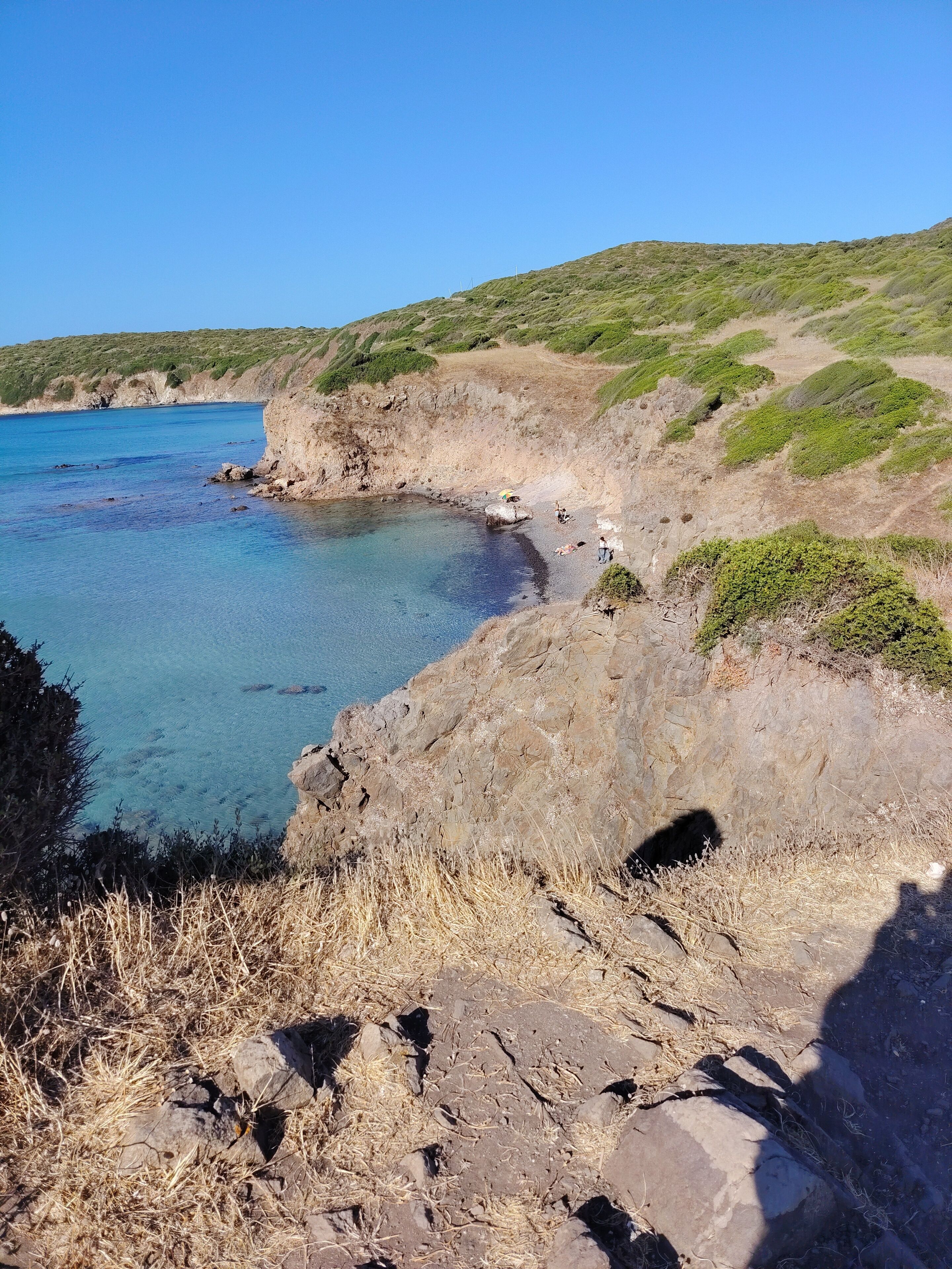 A coastal landscape with a clear blue sea, rocky terrain, and sparse vegetation.