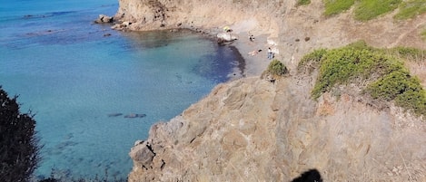 A coastal landscape with a clear blue sea, rocky terrain, and sparse vegetation.