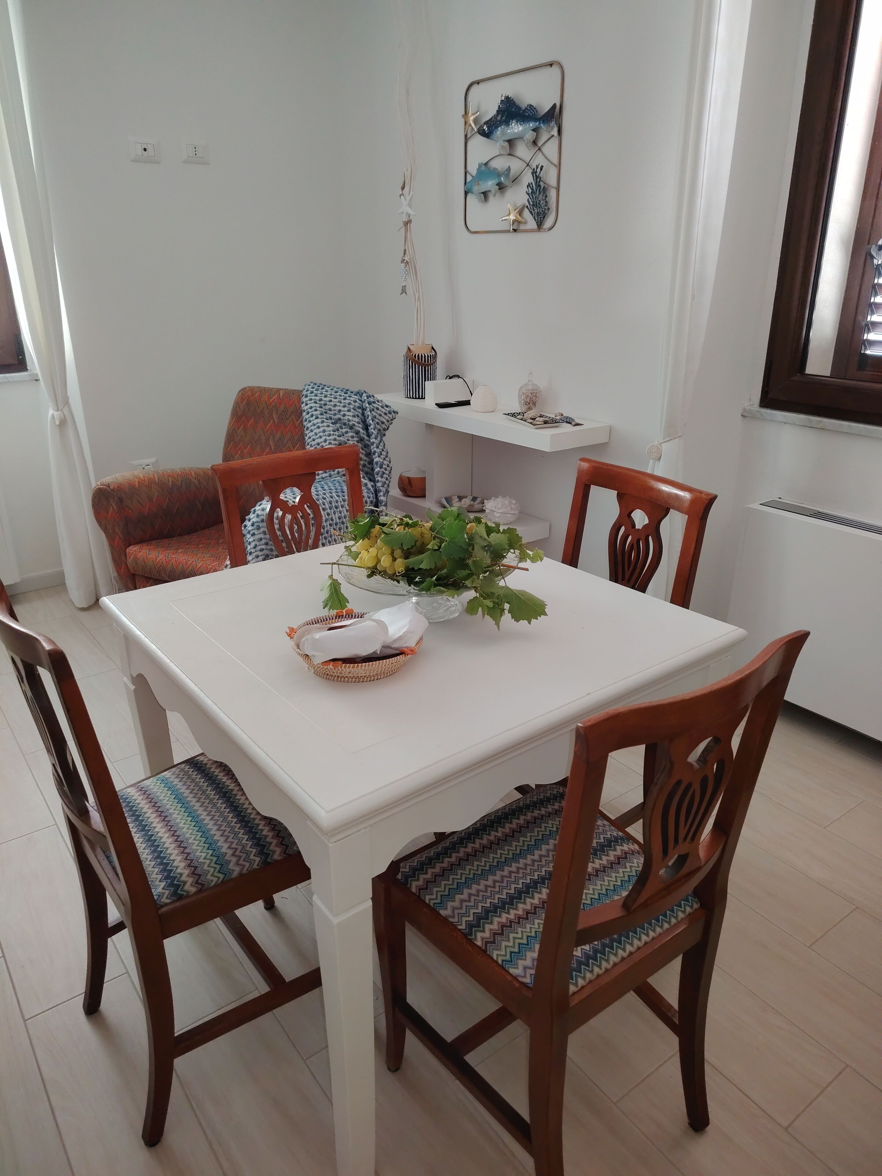A dining area with a white table, wooden chairs with patterned cushions, and a decorative wall piece above the table.