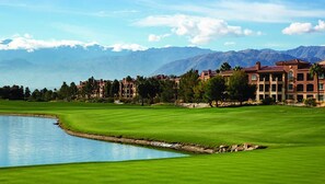 Ein Golfplatz mit Teich, umgeben von Gebäuden vor einer Bergkulisse.