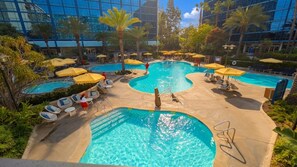 A large indoor swimming pool with multiple lanes, surrounded by lounge chairs and umbrellas, and a backdrop of modern buildings.