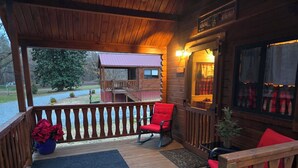 Um deck de madeira com uma cadeira vermelha e uma mesa pequena, um tapete azul, e vista para uma cabana com telhado vermelho.