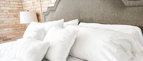 A bed with white bedding and pillows, a grey upholstered headboard, and a brick wall in the background.