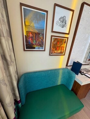A room with a teal sofa, framed artwork on the wall, and a window with patterned curtains.