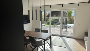 Moderni keittiö, jossa on puinen pöytä ja tuolit, seinälle asennettu televisio sekä liukuovi, joka johtaa ulkotilaterassille.