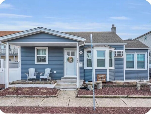 A residential house with a front porch, two white chairs, a small garden area, and a seasonal rental sign.