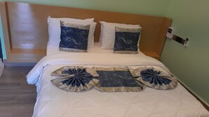 A neatly made bed with a white duvet and decorative pillows, a wooden headboard, and a bedside table with a lamp. - IGAWILO RESIDENCE HOTEL (Dodoma)