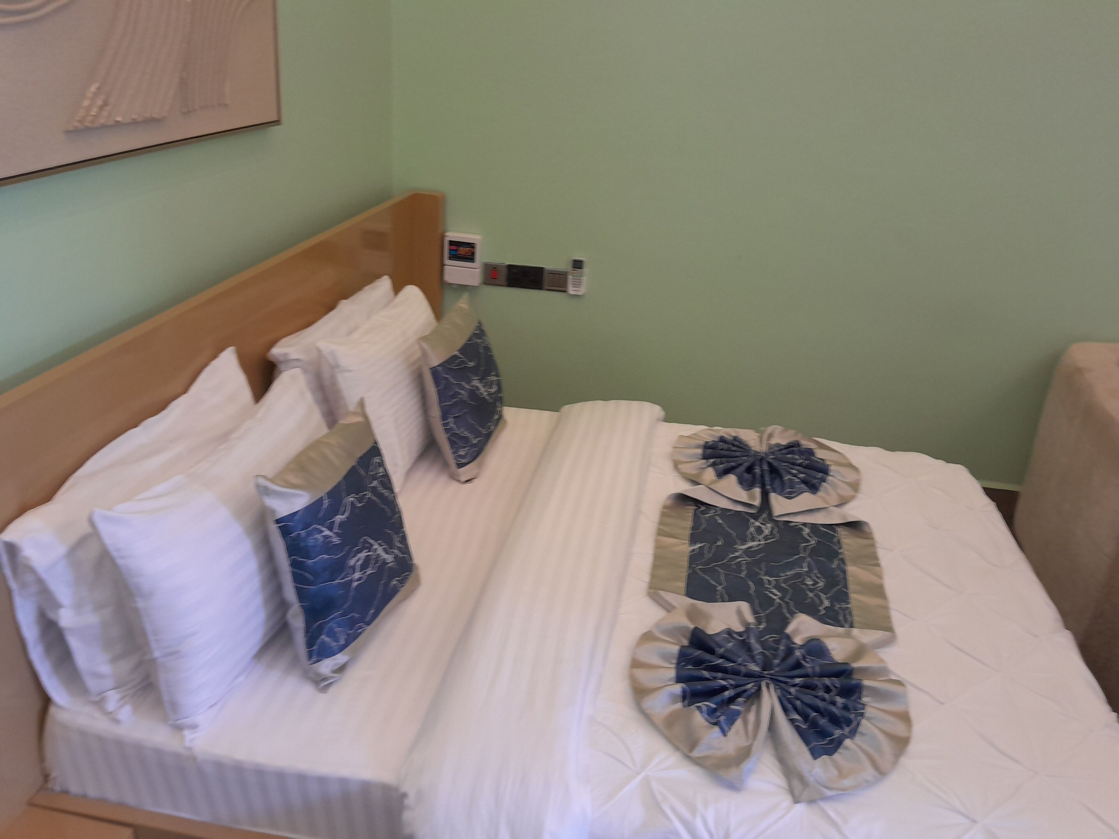 A neatly made bed with white bedding and decorative pillows, a bedside table with a clock, and a wall-mounted thermostat.