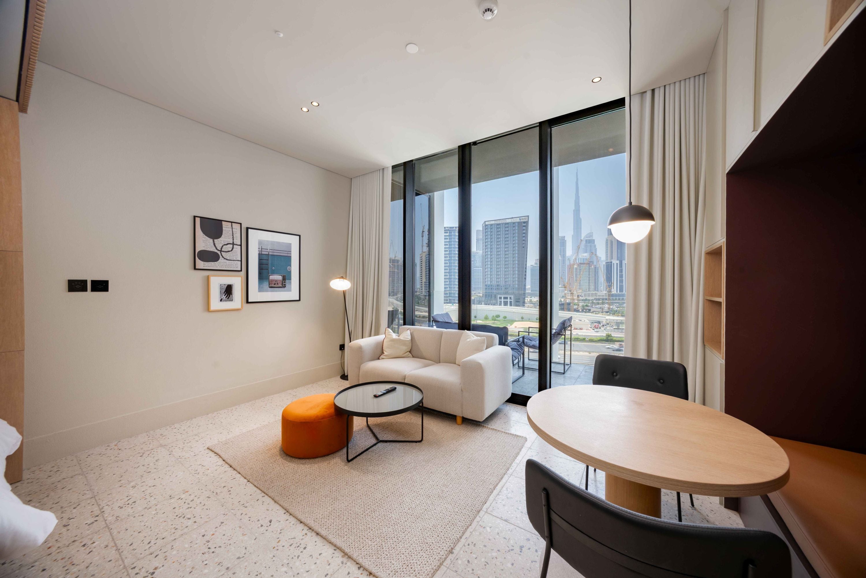 Une chambre d’hôtel moderne avec une grande fenêtre, un canapé, une table ronde et un petit pouf orange.