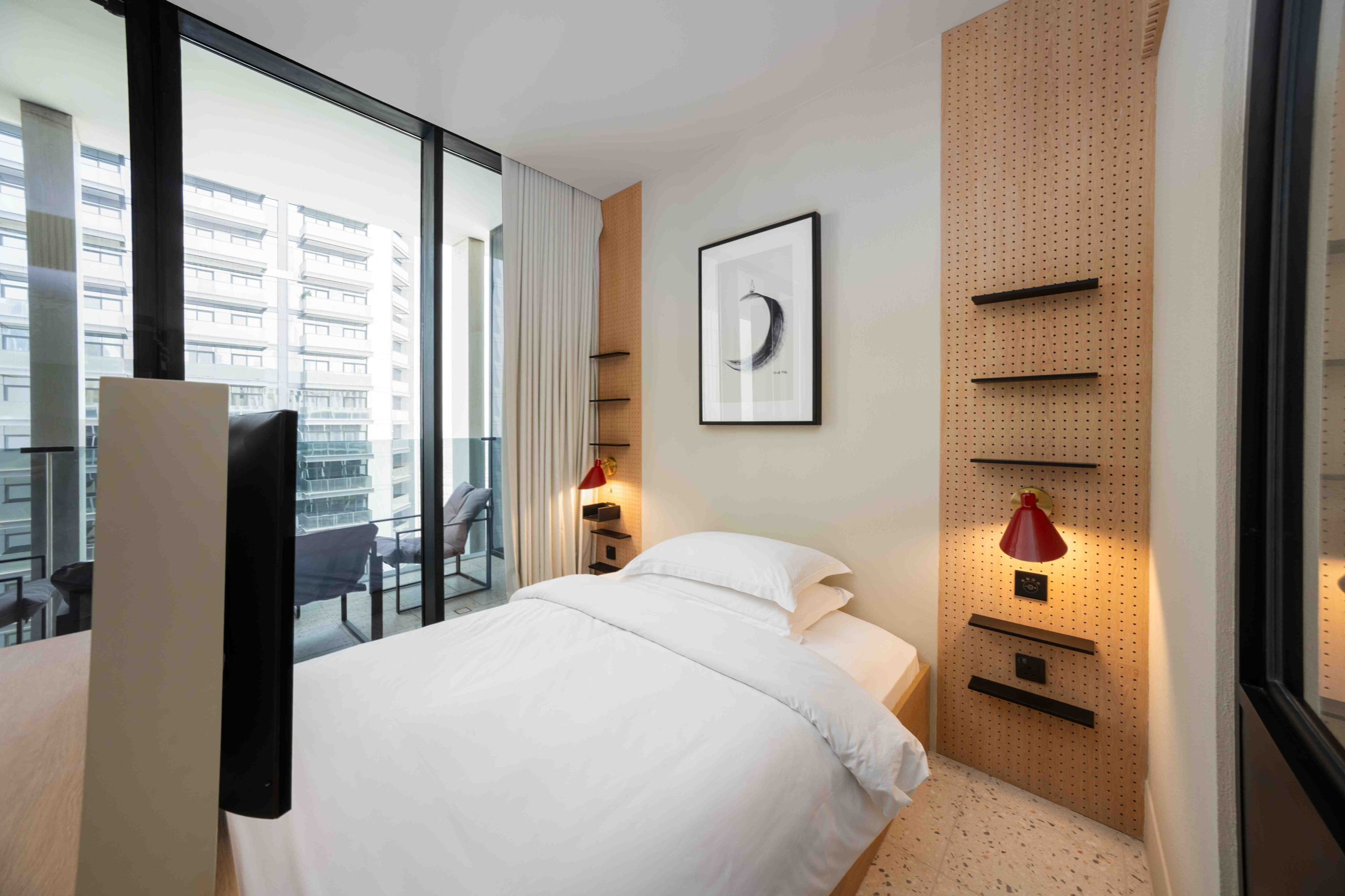 Habitació d'hotel amb un llit, un escriptori, una cadira, una finestra amb vista a edificis i una prestatgeria penjada a la paret amb una làmpada vermella.