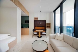 A modern hotel room with a bed, a sofa, a dining area with a table and chairs, and a view of the city through large windows. - Contemporary Studio w/ Burj View (Dubai)