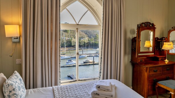 A hotel room with a large bay window, a wooden dresser with a mirror, and a bed with a white comforter. - Riverside Cottage With Terrace (Fowey)
