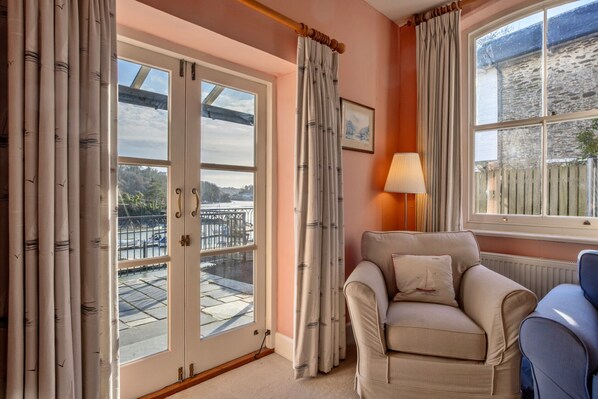 A room with a chair, a lamp, and a view of a dock and water. - Riverside Cottage With Terrace (Fowey)