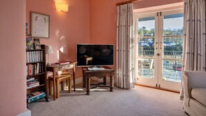 A living room with a television, bookshelves, a small table, and a view of a marina through a glass door. - Riverside Cottage With Terrace (Fowey)