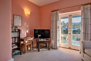 A living room with a television, bookshelves, a small table, and a view of a marina through a glass door. - Riverside Cottage With Terrace (Fowey)