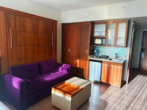 A compact living space with a purple sofa, a wooden coffee table, and a kitchenette with wooden cabinets and a dishwasher.