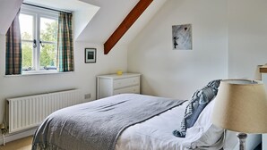 A bedroom with a sloped ceiling, a bed with a grey blanket, a bedside lamp, a dresser, and a window with curtains. - Historic Cornish Valley Cottage for 5 (Crediton)